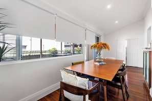 A dining area with a wooden table, chairs, a vase with flowers, and a view through large windows.