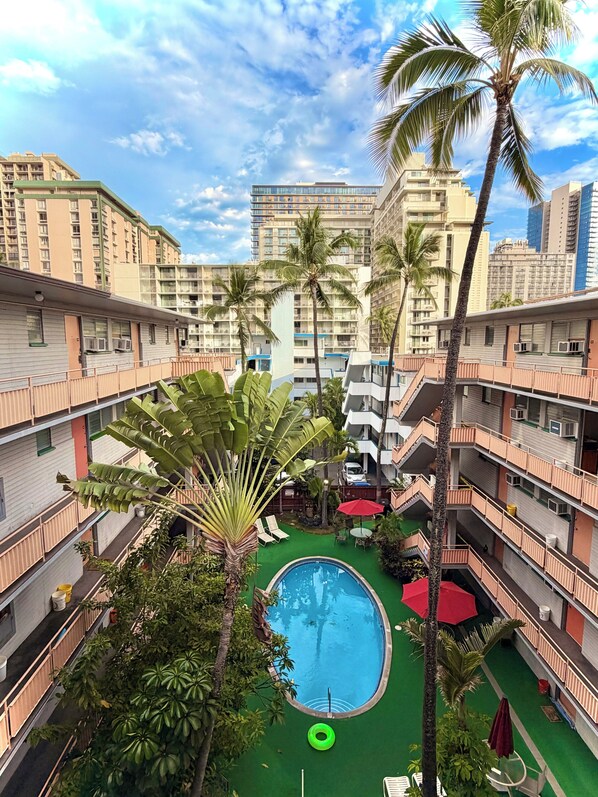 A courtyard with a swimming pool, palm trees, and high-rise buildings in the background. - Waikiki Condo with Full Kitchen 5 min Walk to Beach (Honolulu)