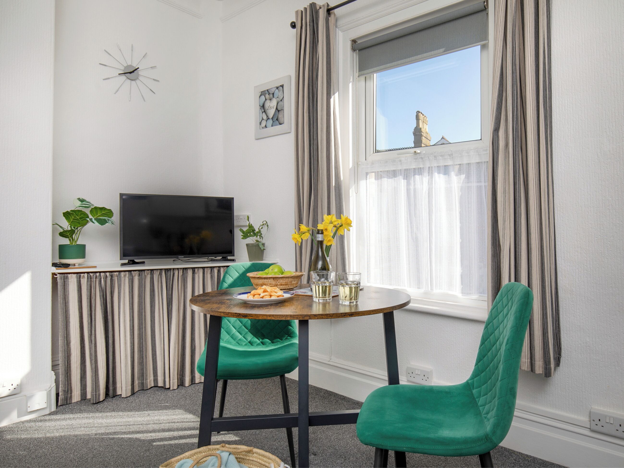 A modern living room with a television, green chairs, a dining table with food, and a window with curtains.