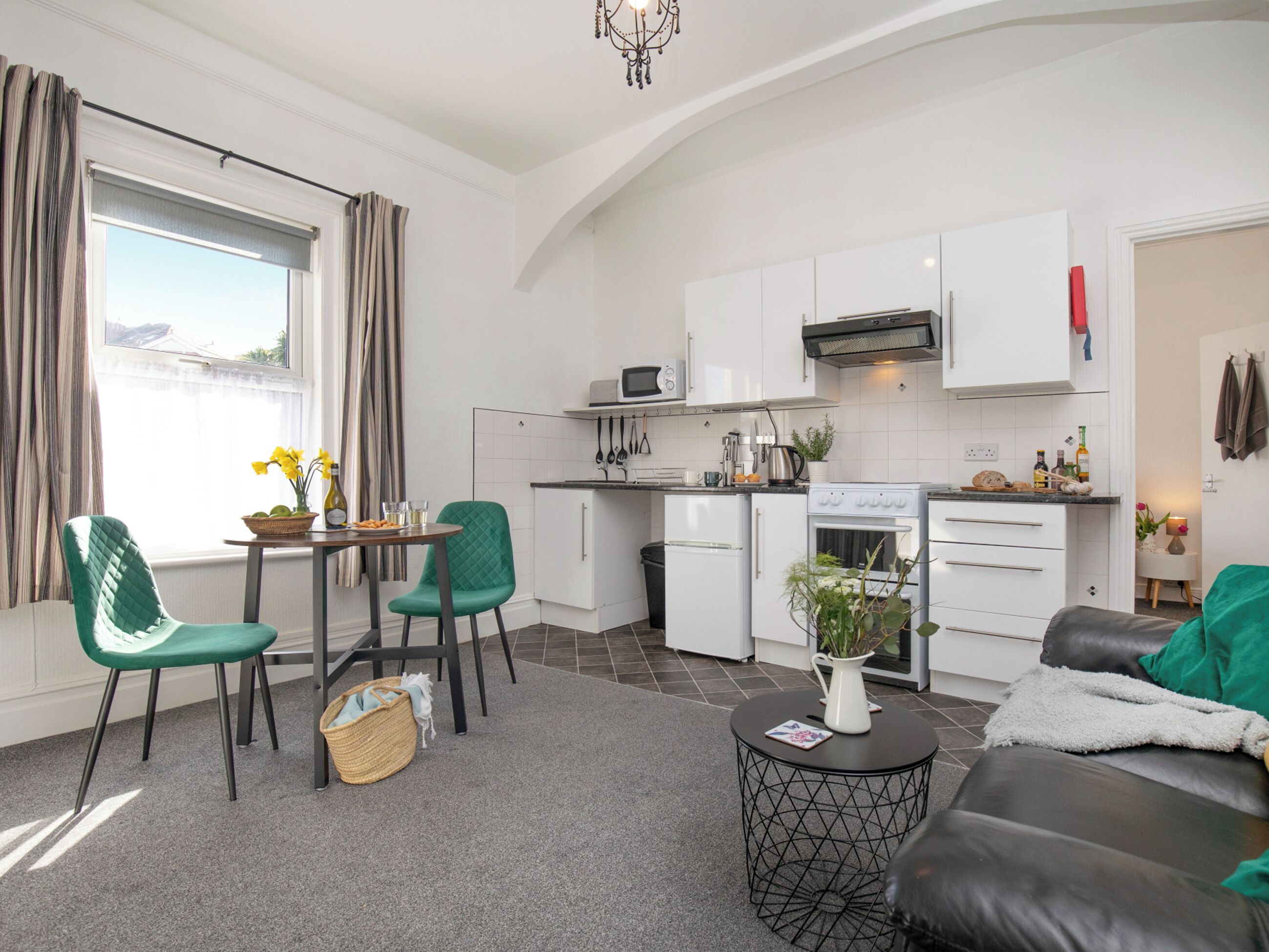 A compact kitchen with white cabinets, a microwave, a stove, and a sink. There is a dining area with a table and chairs, and a living area with a sofa.