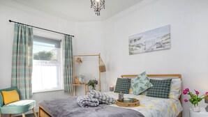 Ein Schlafzimmer mit Bett, Nachttisch, Stuhl, einem Fenster mit Vorhängen, einer Wandablage und einem Gemälde.