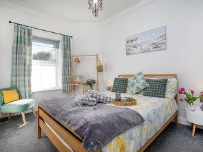 Ein Schlafzimmer mit Bett, Nachttisch, Stuhl, einem Fenster mit Vorhängen, einer Wandablage und einem Gemälde.