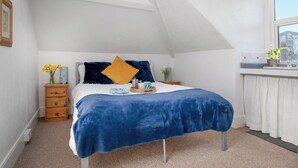 Quarto com teto inclinado, cama com cobre azul, mesa de cabeceira com um vaso de flores e janela com vista para edifícios.
