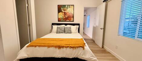 A bedroom with a bed, a painting, and a hallway leading to another room.