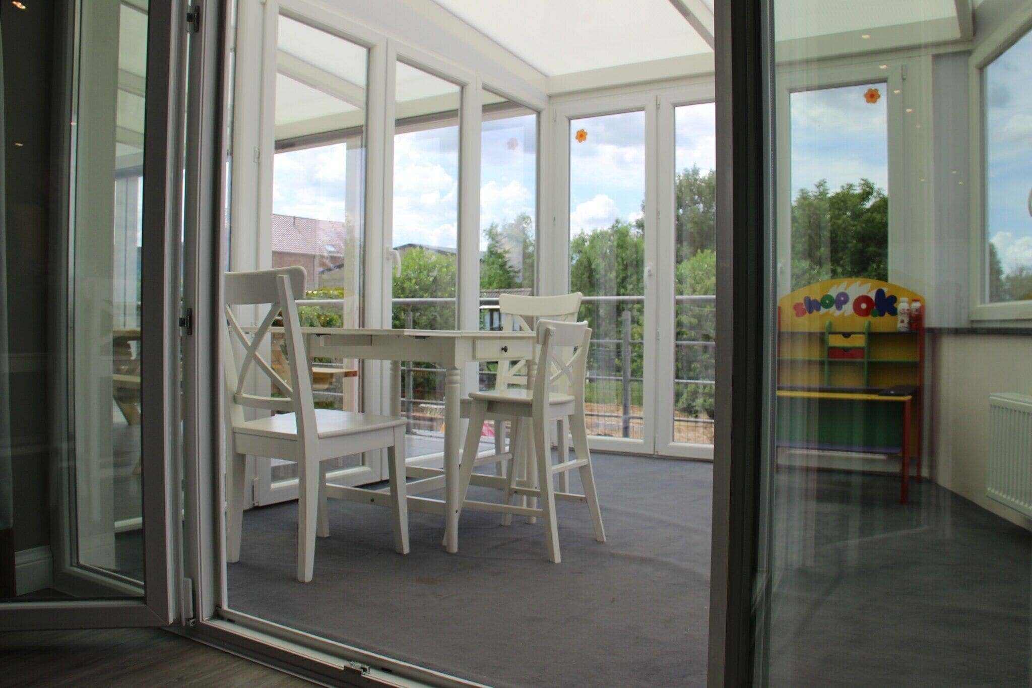 A room with a glass door leading to a balcony, a white table with chairs, and a children's play area with colorful furniture.