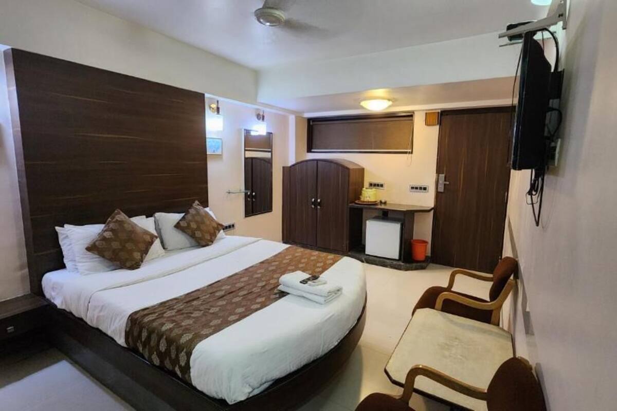 A hotel room with a bed, a desk, a chair, a wardrobe, a television, and a small table with a lamp.