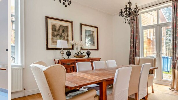 Comedor con mesa de madera, sillas blancas, dos cuadros enmarcados, una araña y ventanas amplias con cortinas.