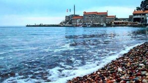 Escena costera con una playa de guijarros, un edificio de techo rojo y una masa de agua.