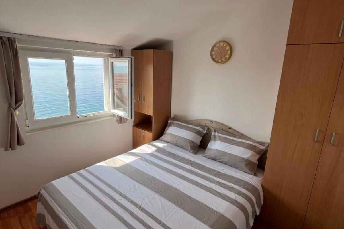 Ein Schlafzimmer mit einem Bett, einem Holzschrank, einem Fenster mit Vorhängen und einer Wanduhr.