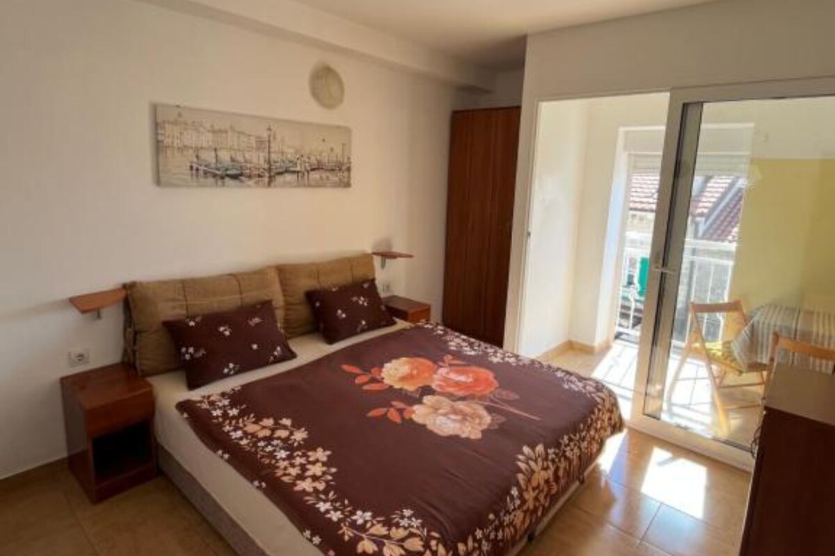 Ein Schlafzimmer mit einem Bett, einem Nachttisch, einer Wanduhr, einer zur Balkontür führenden Tür und einem Wandbild.