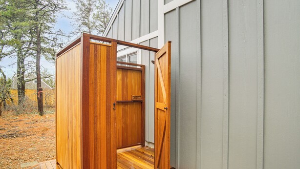 Eine Umkleidekabine für eine Dusche im Freien aus Holz mit Tür, eine Holzterrasse und ein Gebäude mit senkrechten Verkleidungen im Hintergrund.