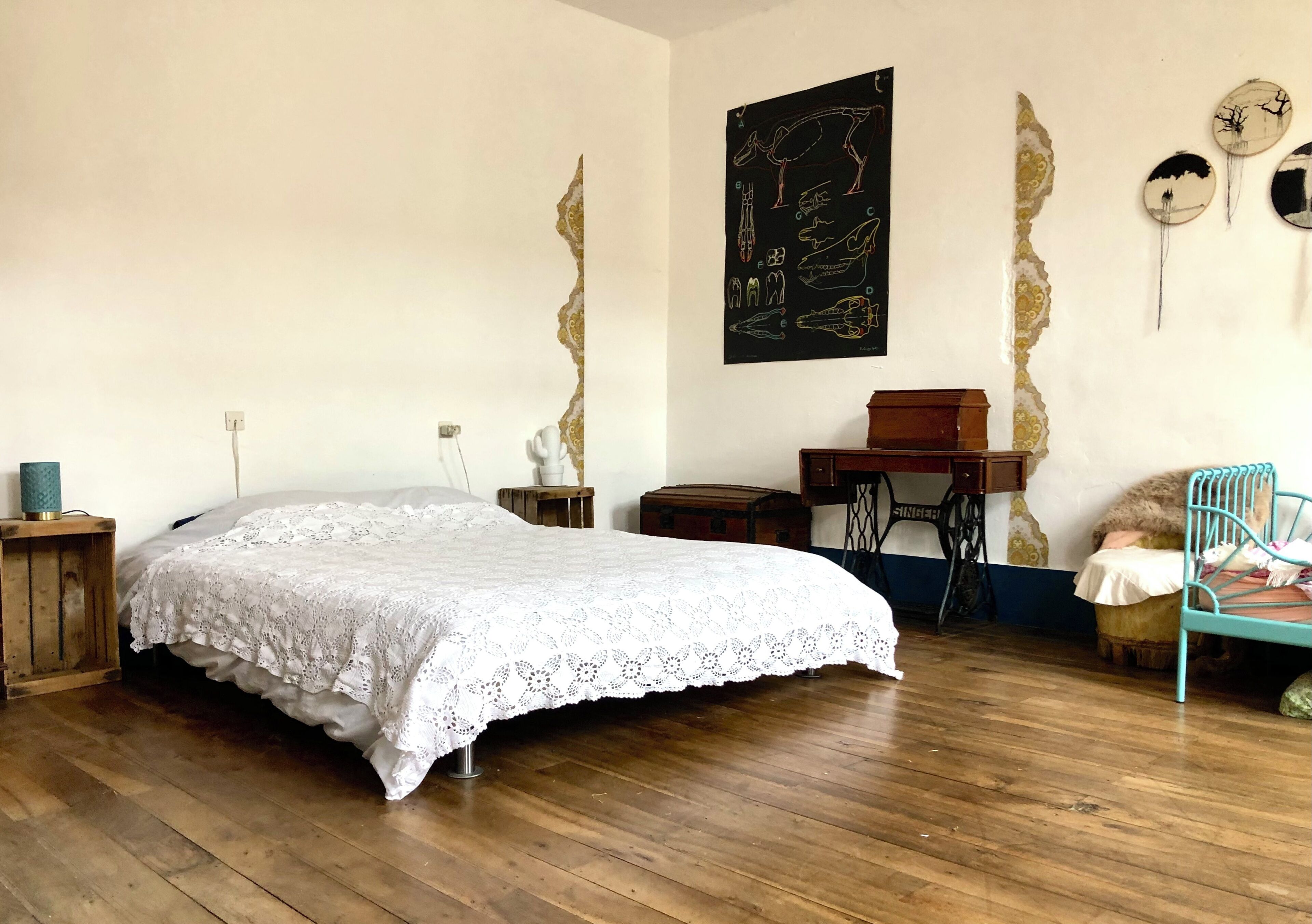 A bedroom with a wooden floor, a bed with a white lace bedspread, a sewing machine, and a chair.