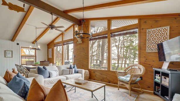 Ein gemütliches Blockhaus-Interieur mit Holzbalkendecke, großen Fenstern, einer Couch mit Kissen, einem Couchtisch und einem Korbstuhl.