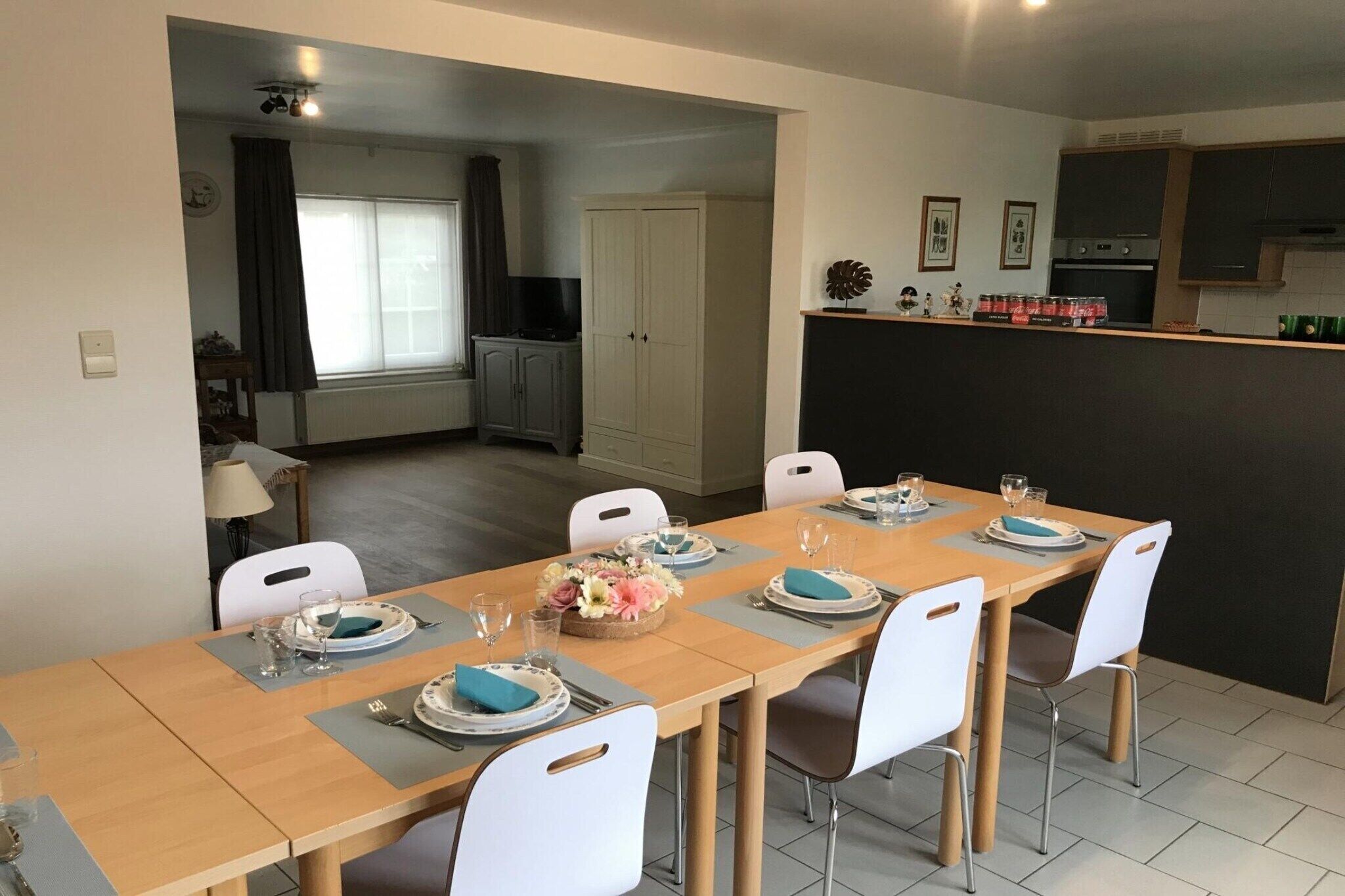 A dining area with a long table set for six, white chairs, and a kitchen area with cabinets and a countertop.