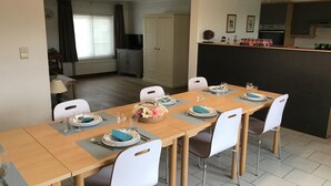 En matplats med ett långt bord som är dukat för sex personer, vita stolar och ett köksområde med skåp och en bänkskiva.