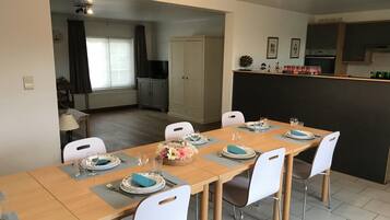 A dining area with a long table set for six, white chairs, and a kitchen area with cabinets and a countertop.