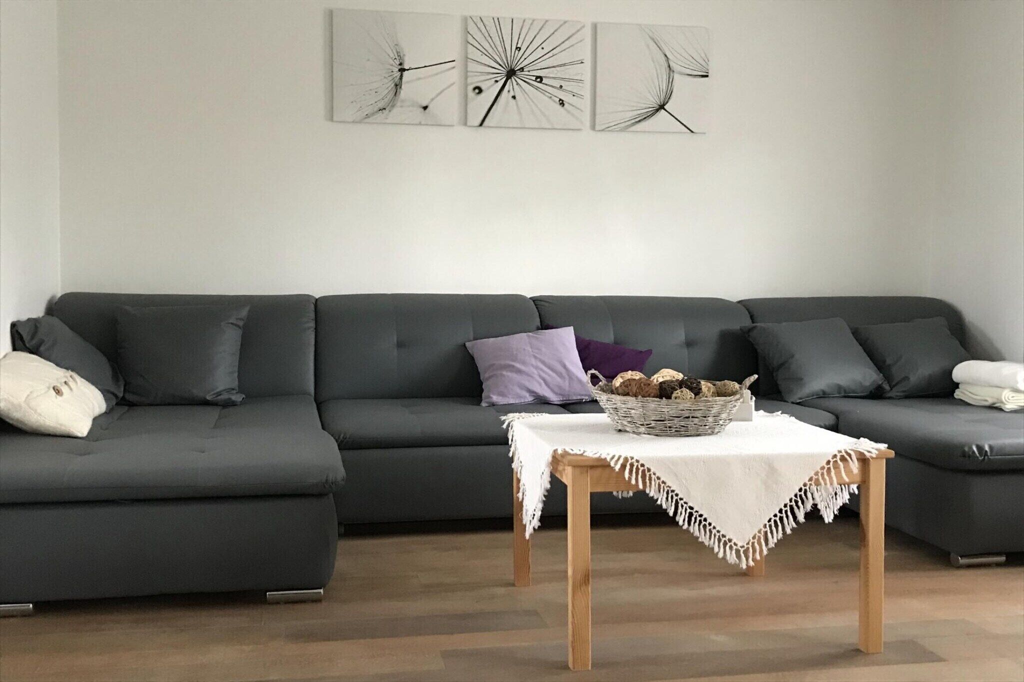 A modern living room with a dark grey sectional sofa, a small wooden table with a basket of bread, and three abstract wall art pieces.