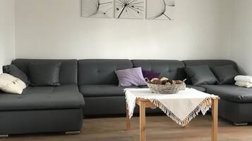 A modern living room with a dark grey sectional sofa, a small wooden table with a basket of bread, and three abstract wall art pieces.