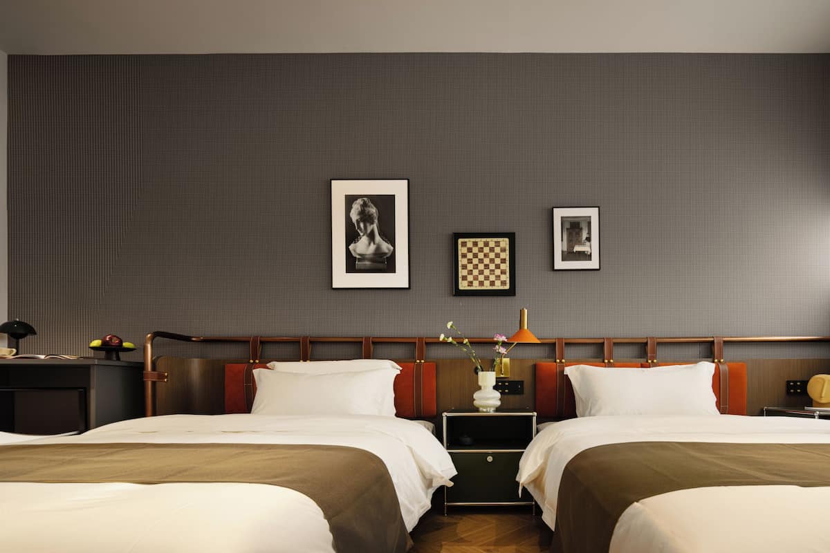 A hotel room with two beds, a desk with a chair, a TV, and framed pictures on the wall.