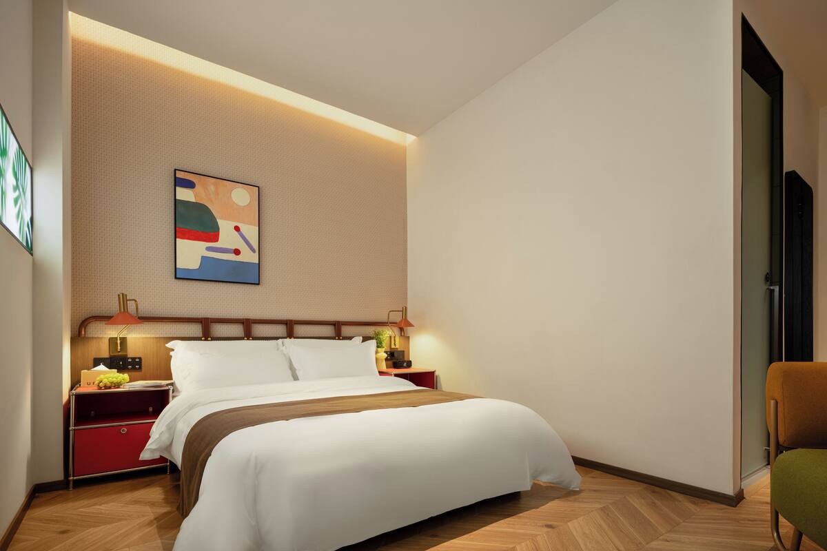 A modern hotel room with a bed, nightstands, a wall-mounted lamp, and a painting.