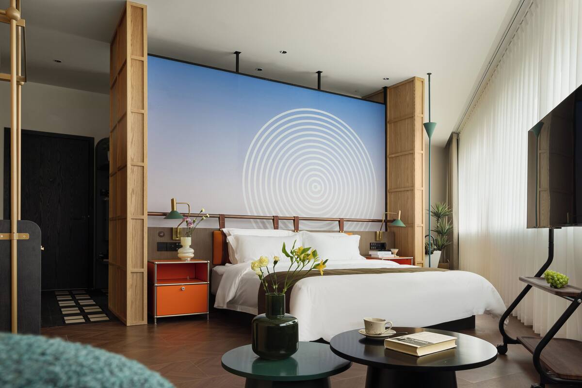 A modern hotel room with a large bed, a bedside table, a nightstand, a wall with a circular design, and a large window.
