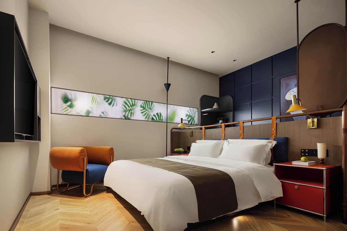 A modern hotel room with a bed, a chair, a bedside table, a lamp, and a wall-mounted TV.