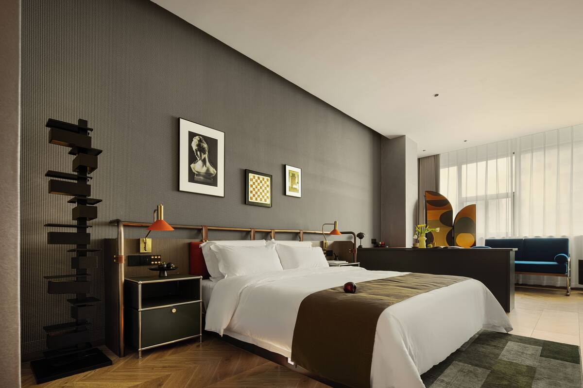 A modern hotel room with a large bed, a desk, a chair, a sofa, and a large window.