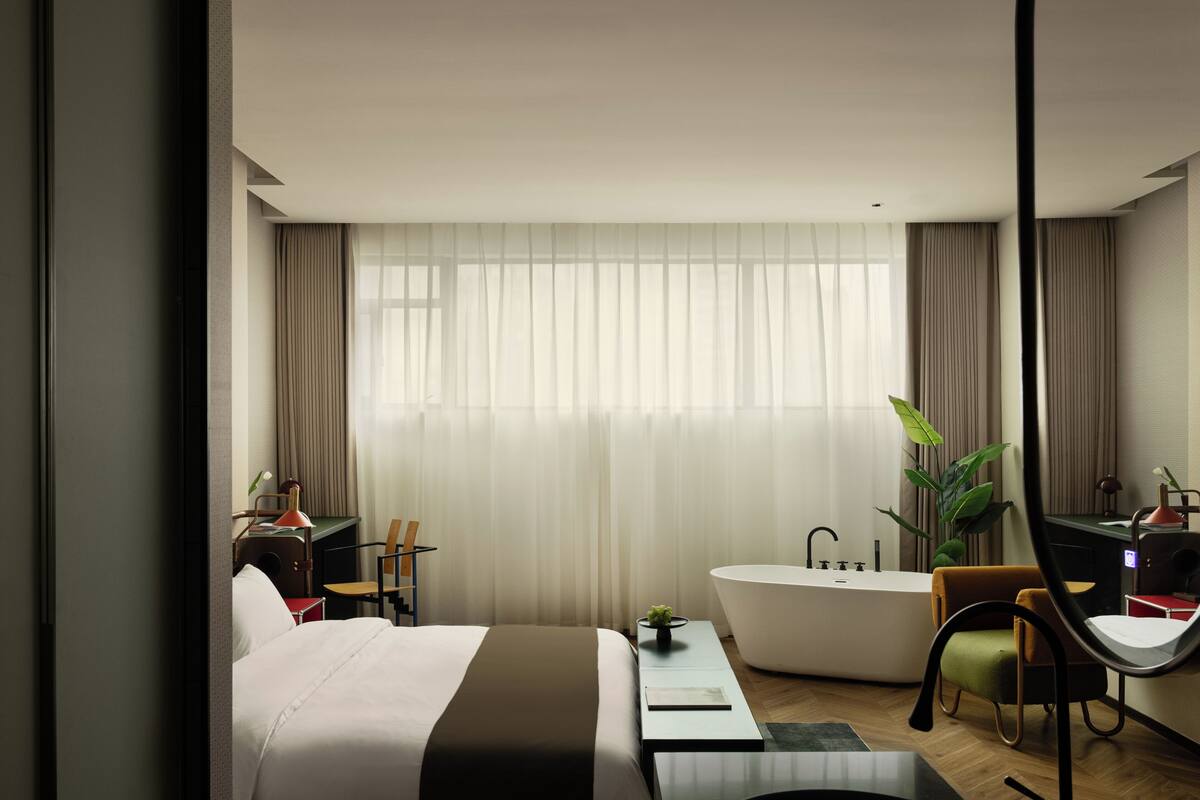 A modern hotel room with a large bed, a sitting area with a desk and chair, a bathtub, and a large window with curtains.