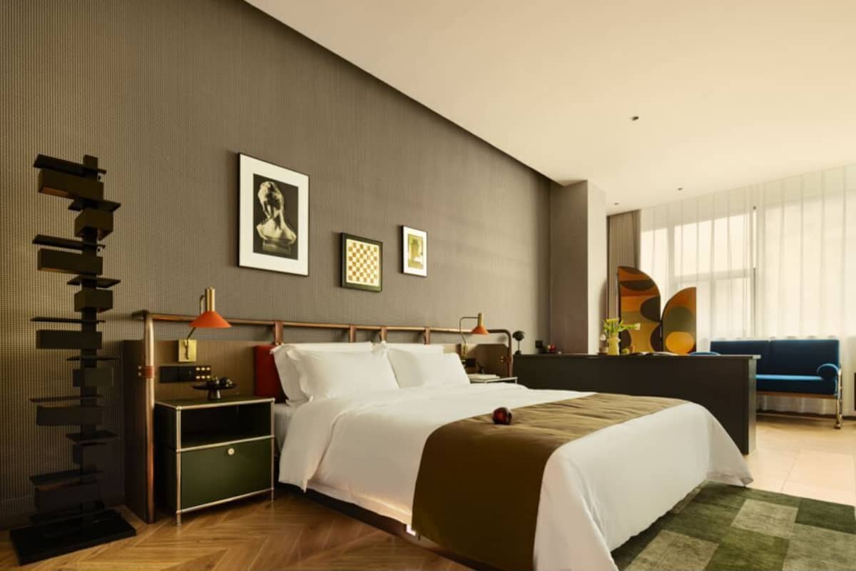 A modern hotel room with a bed, bedside tables, a desk, and a large window.