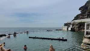 Ein Schwimmbecken mit klarem Wasser, eine steile Felswand und eine Treppe zu einem Gebäude.