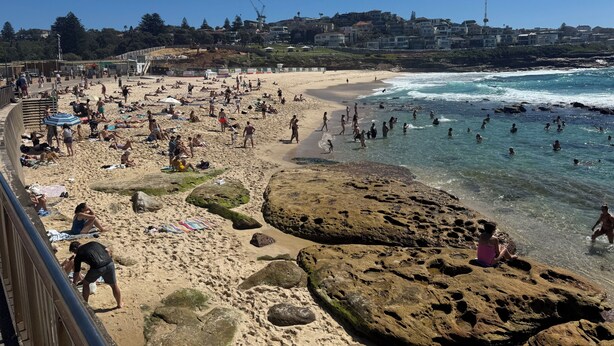 Ein Strand mit badenden und sonnenbäckernden Personen, felsigen Abschnitten und Gebäuden im Hintergrund.