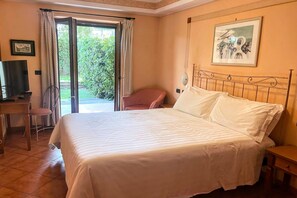 A hotel room with a bed, a desk, a chair, and a view of a garden through a glass door. - Elite Residence Near Rome and Tivoli (Roma)