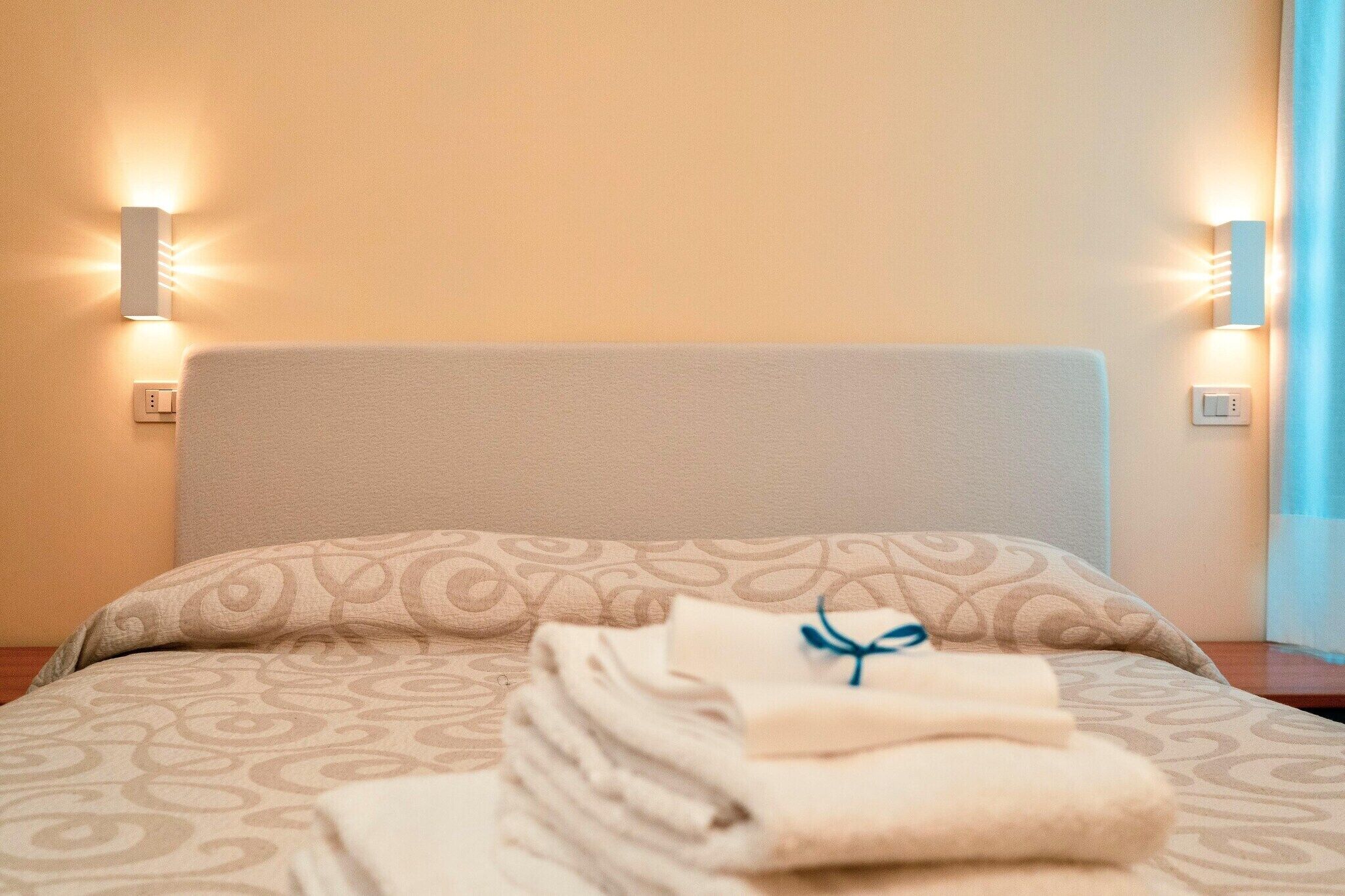 Un lit bien fait, avec une couvre-lit à motifs et des oreillers, une pile de serviettes pliées, maintenue par un ruban bleu, et deux appliques murales de chaque côté du lit.
