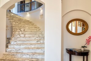 Eine Marmortreppe führt zu einer zweiten Etage, an der Wand hängt ein runder Spiegel und ein kleiner Tisch trägt einen Blumentopf und Bücher.