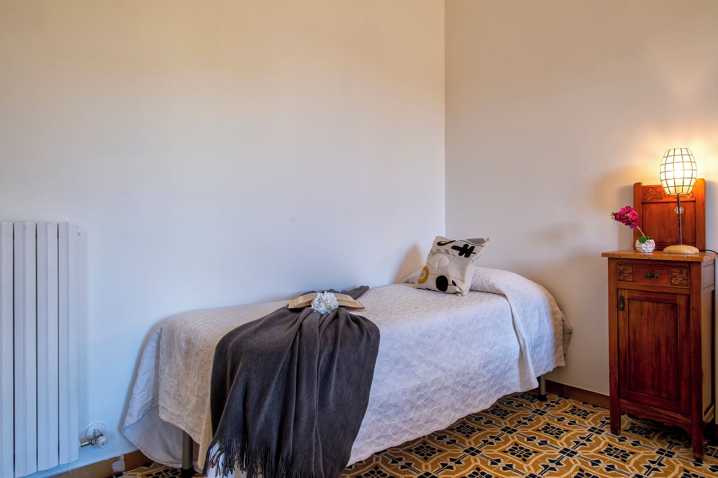 A bedroom with a bed, a radiator, a wooden cabinet, and a wall-mounted lamp.
