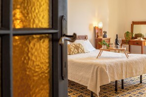 Ein Bett mit gemustertem Überwurf, ein Nachttisch mit Lampe, ein Spiegel und eine Kommode mit Pflanze.