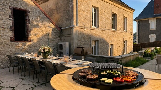 Un espace extérieur aménagé pour les repas, avec une table ronde, des chaises et un barbecue où l’on prépare les aliments.