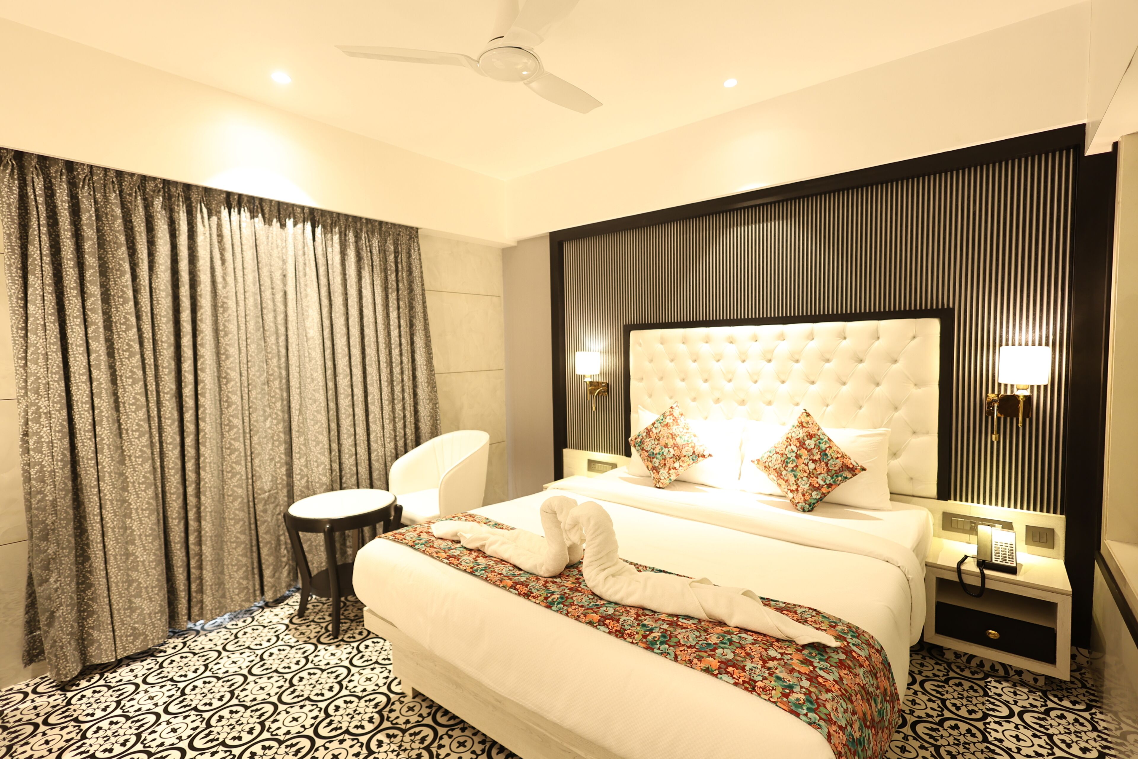A hotel room with a bed, a chair, a small table, a nightstand with a phone, and a ceiling fan.