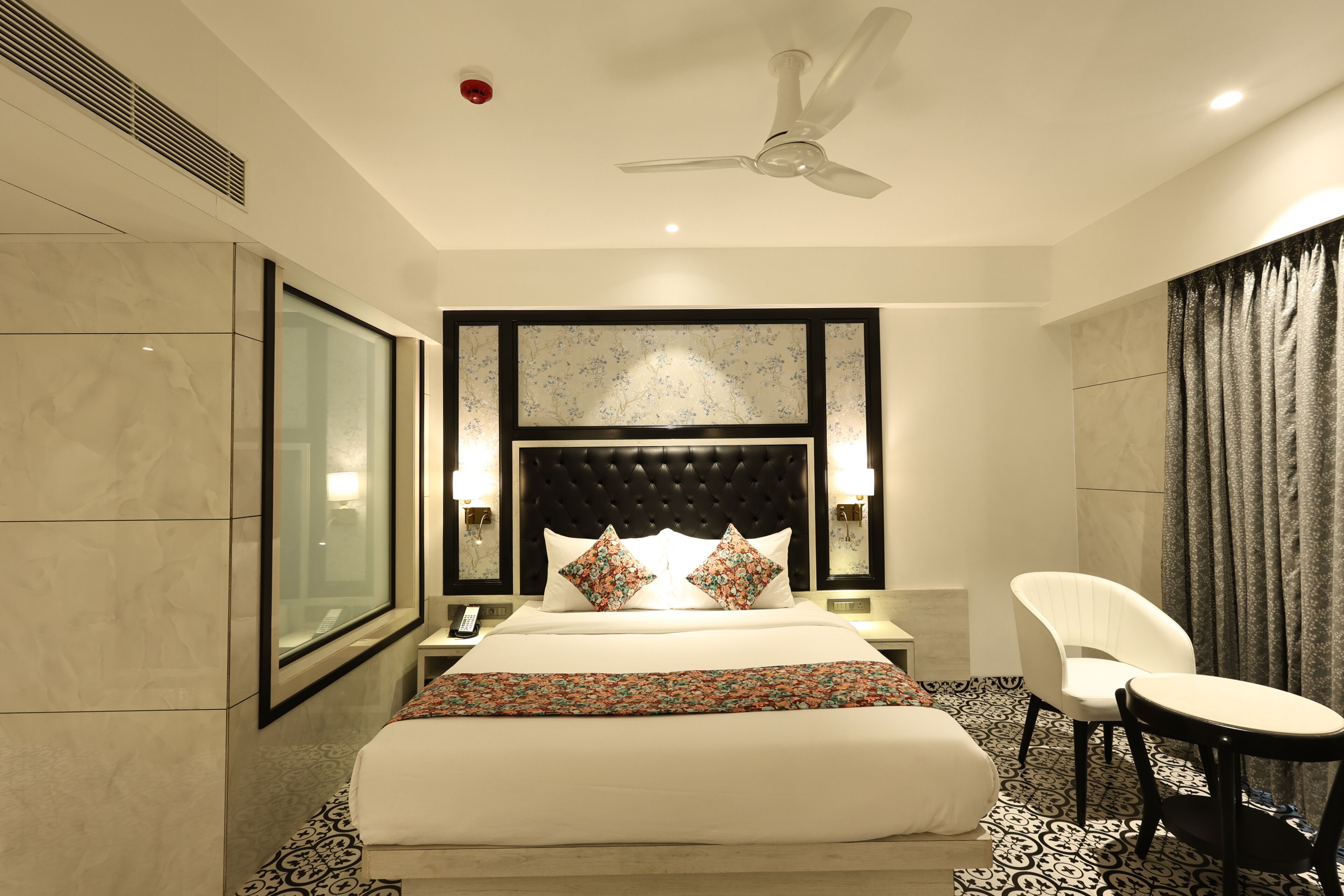 A hotel room with a large bed, a ceiling fan, a nightstand, a chair, and a large window.