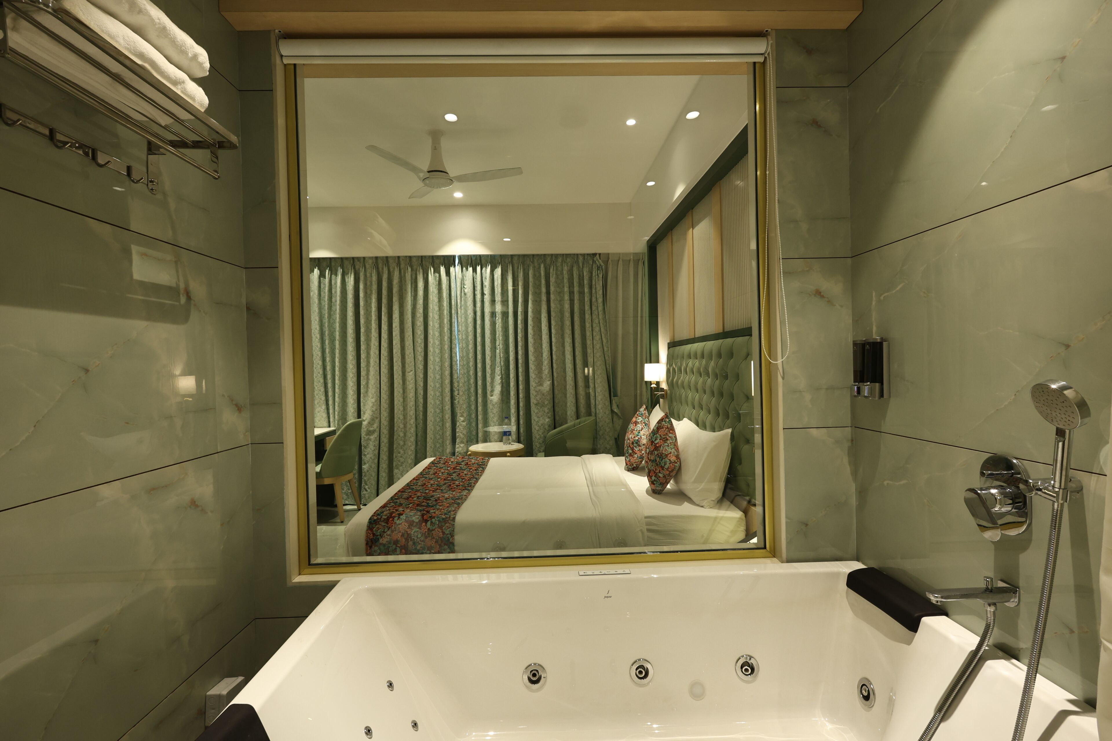 A modern bathroom with a large bathtub, a shower, and a mirror reflecting a bedroom with a bed and curtains.