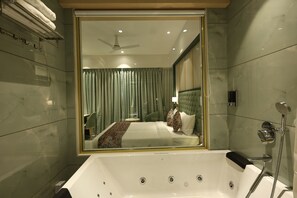 A modern bathroom with a large bathtub, a shower, and a mirror reflecting a bedroom with a bed and curtains. - SiO Suites (Navi Mumbai)