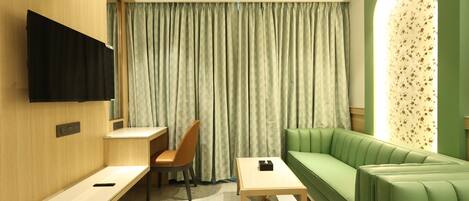 A hotel room with a green sofa, a desk with a chair, a TV mounted on the wall, and a window with curtains.