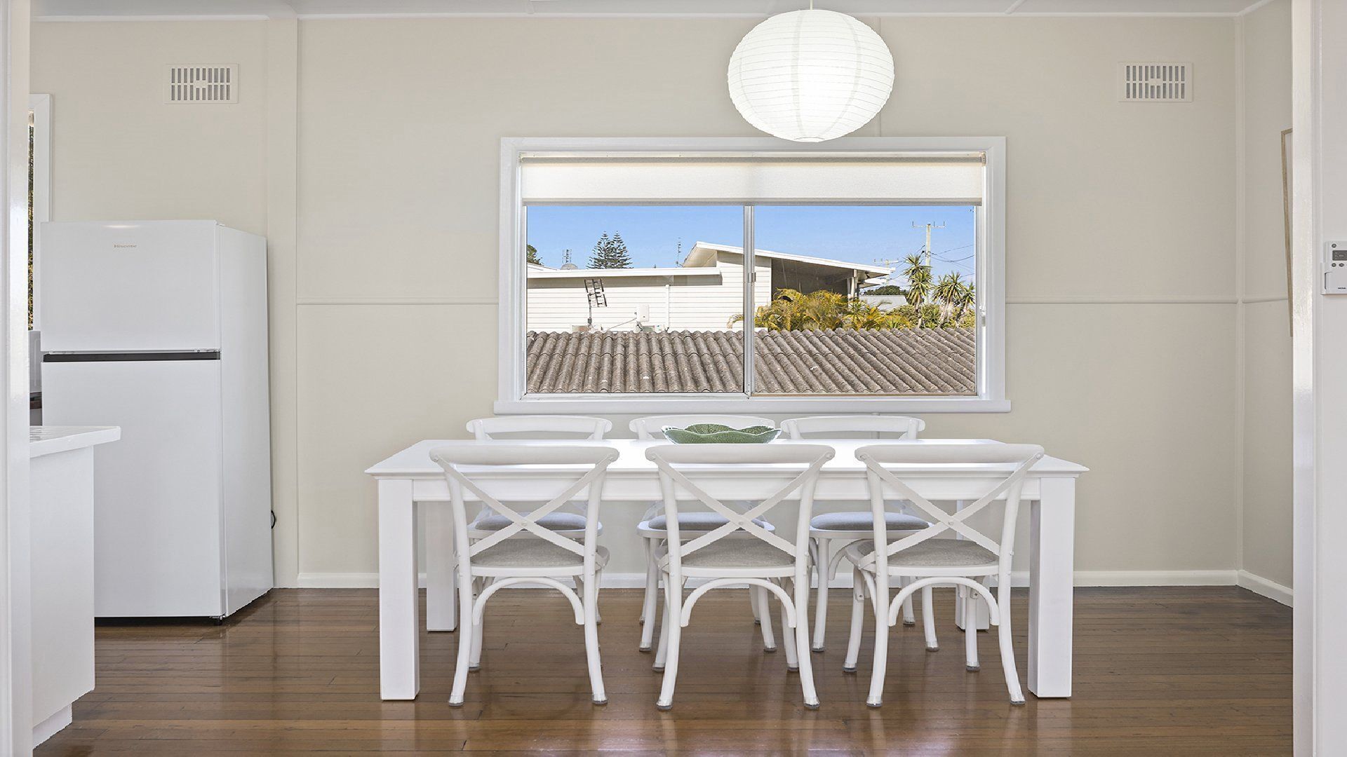 Ein Essbereich mit Tisch und Stühlen, ein Fenster mit Blick auf ein Gebäude und Bäume sowie ein weißer Kühlschrank.