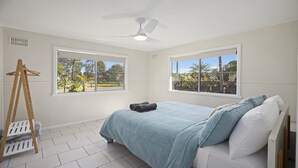 Quarto com cama, ventilador de teto e duas janelas com vista para o verde.