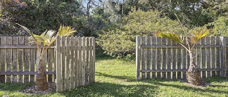 Una cerca de madera con un área cubierta de césped al frente, rodeada de árboles y arbustos.