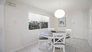 Ein Essbereich mit einem runden Tisch, weißen Stühlen, einem Fenster mit Jalousien, einer Deckenleuchte und einer Tür.