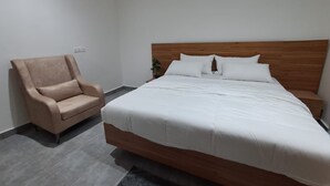 A modern hotel room with a large bed, a wooden headboard, a beige armchair, and a small table with a plant.