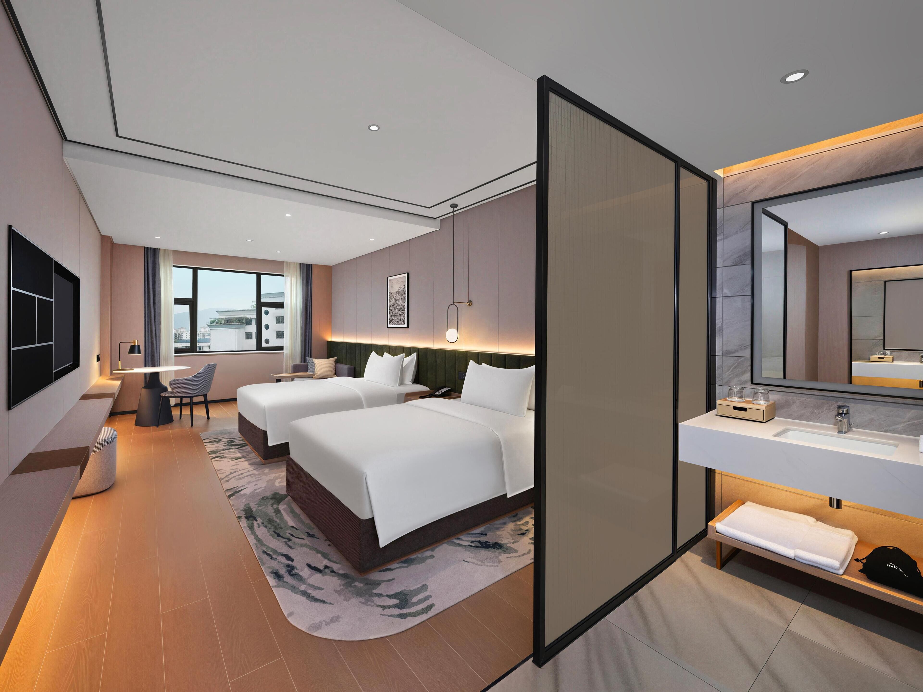 A modern hotel room with two beds, a desk, a chair, a large window, a bathroom with a mirror and sink, and a view of the city.