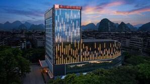 Een modern hotel met een glazen gevel en verticale lichtaccenten, omgeven door weelderige begroeiing en bergen op de achtergrond.