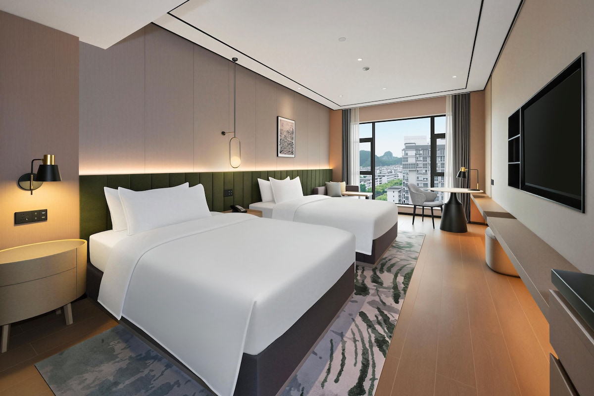 Ett modernt hotellrum med två sängar, ett skrivbord, en stol, en TV och utsikt över staden.